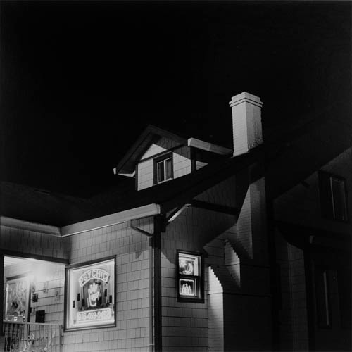 A long exposure photograph of a house with neon signs in the window. The neon lights show a phone number, the symbol of a crescent moon, candles, and the word "Psychic". The photo is shot at an angle showing the corner of the house with the front to the left and a side to the right. The dark pitched roof can barely be made out against the black of the night sky. A small attic room window can be seen on top with a chimney protruding to the right.