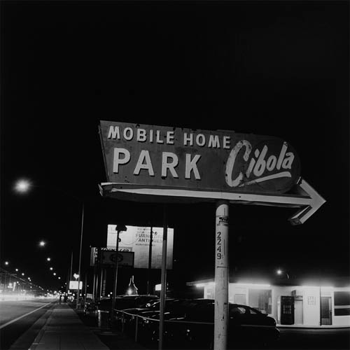 A long exposure photograph of a large sign that reads "Mobile Home Park Cibola". The words mobile, home, and park are in capital letters while the word Cibola is in a script font and is written at an angle with a swooping stroke underneath. The sign also features an arrow pointing to the right. To the left of the sign the glow from the street lights and traffic can be seen. The sign itself features no illumination.