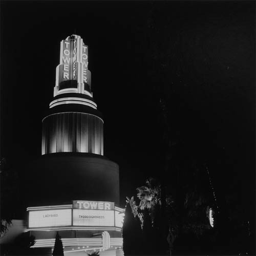 A long exposure photograph of a tower shaped building. The building includes a movie theater marquee on the front, below the tower. The top of the tower features two bisecting rectangular structures making a plus or x if viewed from the top. Below the plus structure the tower become cylindrical. Each face of the rectangle has the word "tower" shown in neon lights. The word is displayed vertically in all capital letters. The edges of the rectangular structures are lined with thin neon lights down the length. Where the tower becomes cylindrical, the neon lines go around the circumference horizontally. The movies listed on the marque are "Ladybird" and "Thoroughbreds". In the foreground palm trees can be seen along with faint outlines of other trees.