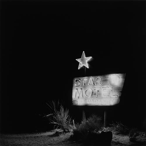 A long exposure photograph of a neon sign. The sign reads "Star Motel", in the upper right corner the letters "TV" appear in a circle. At the bottom more neon lights read "No Vacancy". Floating above the main portion of the sign a neon lit star hangs in the darkness. The structure that attaches the star to the rest of the sign can just barely be seen. The sign is isolated in the darkness with some desert shrubs illuminated by the neon in the foreground.