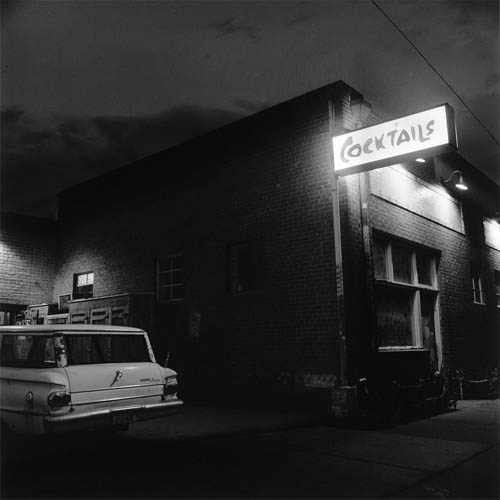 A long exposure photograph of the exterior of a cocktail lounge. Next to the building is an old station wagon, only the rear end is visible. Hanging off the top od the building is a brightly lit sign that reads "Cocktails", it is illuminating the bricks of the building. Boarded up windows below the sign are lit up by additional lights. behind the car an assortment of palets and other objects can be seen.