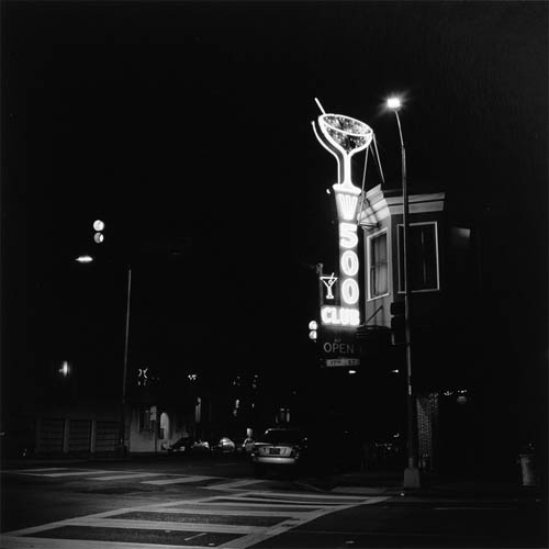 A long exposure photograph of a bar with a vertical neon lit sign that reads "500 Club" the sign features cocktail glasses in addition to the text. The bar and sign are on the right side of the photograph and in the foreground is a crosswalk illuminated by the street lights. To the left is the street faded in to the darkness.