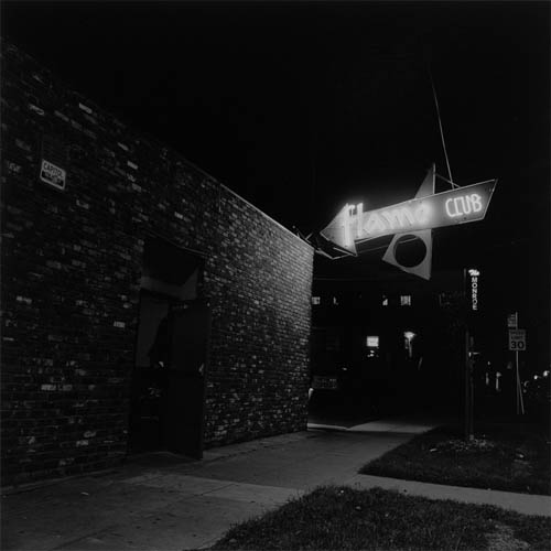 A long exposure photograph of a bar with a googie style neon sign that reads "Flame Club". The sign is illuminating the brick work of an otherwise featureless short rectangular building. The door is propped open and you can just catch a glimpse of the mystery inside.