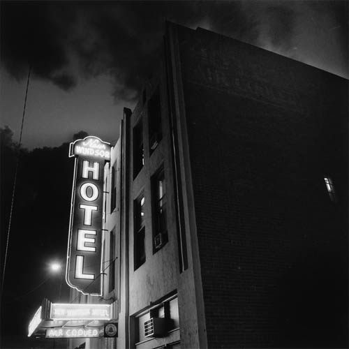 A long exposure photograph of a hotel taken at the corner of the building. The front has a large vertical neon sign that spans to the top of the building. The sign reads "New Windsor" at the top horizontally, the word "Hotel" is vertical and runs the length of the sign. At the bottom it reads "Air Conditioned" horizontally. The side of the building shows the brickwork with a faded painted sign the reads "Hotel Air Conditioned". One small window is on the side of the building, the light is on and you can just barely make out the interior. In the sky monsoon clouds can be seen in the dark sky.