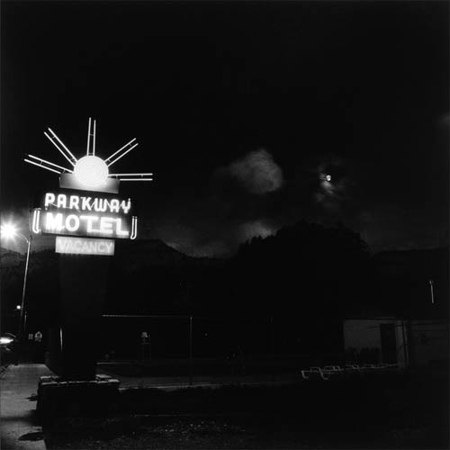 A long exposure photograph of a roadside motel. To the left of the frame, the highway is visible and a neon sign sits right on the edge. The sign is brightly lit, the top of the sign is modeled off of the sun with a circle featuring concentric tubes of lit neon. Five lines protrude at angles from the center to mimic sun rays. The bottom of the sign reads "Parkway Motel Vacancy" with each word on one line. To the right of the sign a fence can be just barley seen in the dark foreground, behind it is the motel pool. In the dark sky above clouds can be seen crossing the full moon. In the background the dark outline of a mountain can be seen against the slightly less dark sky.