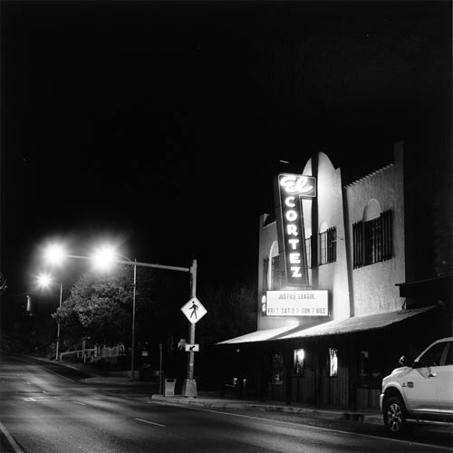 A long exposure photograph of a 1930s movie theater. The theater features an impressive marque and neon sign. The neon sign is vertical and reaches to the top of the building. At the very top the word "El" is horizontal coming out from the face of the building perpendicular to the front of the building. The sign then goes down at a slight angle and the word "Cortez" extends the length vertically. At the bottom is the marque, the movie listed is "Justice League". To the left of the frame the street can be seen. There is a street light extending over the road, its lights are illuminating the wet road. A cross walk sign is seen on the pole. The road extends into darkness in the background. In the foreground a pickup truck can be seen parked on the road.