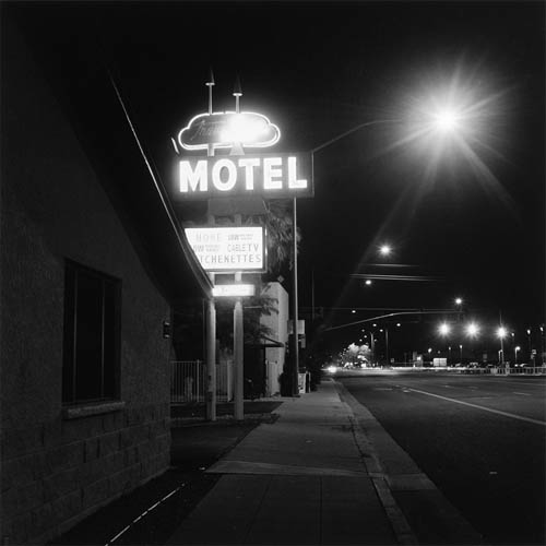 A long exposure photograph of road side motel sign. On the left side of the frame in the foreground a darkened building can be seen. At the end of the building the sign is lit up. At the very top of the sign a bubble shape with the letters "Trava" is visible, the rest of the words are too blown out with light to be legible. Below that the word "MOTEL" is horizontal in all caps. At the bottom of the sign is a marque that advertises "Phone", "Low vacation rates", "Low monthly rates", "Cable TV", and "Kitchenettes". In the middle of the frame a sidewalk divides the sign and building and the street to the right. Street lights can be seen down the distance of the street.