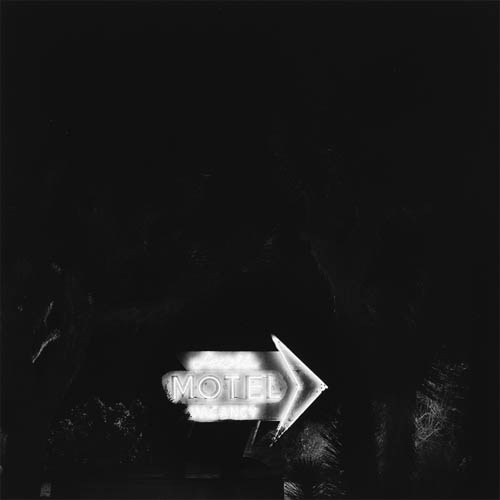 A long exposure photograph of a motel sign in the middle of the desert. In the foreground the faint dark outline of joshua trees can be barely seen. Behind the trees in the middle of the frame the brightly lit sign can be seen. Some desert shrubs in between the sign and the trees are illuminated by the neon lights. The sign is in the shape of an arrow pointing to the right. The word "Lewis" is at the top in a script typeface, in the middle the word "MOTEL" is all in caps and is twice as big as the text above, below that is the word "VACANCY" also all caps but in a thin set typeface.