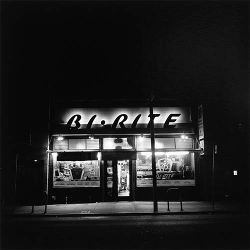 A long exposure photograph of a small neighborhood market. The store appears to be closed at this late hour in the night. The interior lights are on and you can make out the stocked shelves of goods glowing in the windows. Above the windows are giant letters creating the word "Bi-Rite" these letters have lights behind them creating stark contrast between them and the tiled wall behind them. The lights fully illuminate the gleaming tiles. In the foreground the empty sidewalk and edge of the road can be seen.