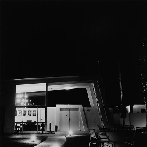 A long exposure photograph of the exterior of a building. The building has a very angular mid-century architecture style. The roof extends beyond the walls containing the building at an angle. It is then bisected by a support structure that creates a slightly obtuse angle. Under the roof the gate of an exterior wall mimics the same angled shape. A large glass window of the building can be seen with the interior illuminated by an atomic-age chandelier. A desk can be seen along with framed art on the walls. A lighted sidewalk path can be seen in the foreground leading to the gate. In the background faint outlines of palm trees can be seen.