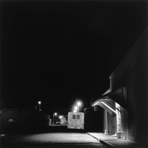 A long exposure photograph of a desolate street. In the center of the frame, in the mid-ground there is a box truck parked facing away from the frame. The aluminum paneling of the rear of the truck is brightly lit by the streetlights. In the right of the frame in the foreground the face of a building can be seen, the building has and awning above a door. A light under the awning fully illuminates the doorway against the dark building. In the background you can see the street being sporadically illuminated by more street lights into the distance.