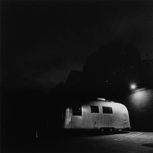 A long exposure photograph of an travel trailer in an empty parking lot. The side of the trailer is facing the frame at a slight angle. There is a brick wall at the edge of the lot that the trailer is backed up to. A single light on the wall is illuminating the riveted aluminum side of the trailer. In the background, beyond the wall, the faint outline of a tree and house can be seen against the black sky.