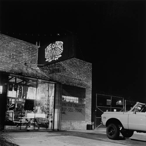 A long exposure photograph of an industrial building that is being used as a custom motorcycle shop. A metal grate is covering the open shop door. The light is on in the building showing the interior of merchandise and old motorcycle parts. The front of the building has exposed brickwork, painted on the bricks are the words "Vintage Choppers" & "Buy, Sell, Trade". There is a sign hanging on the front of the building that reads "Love Cycles" and has a logo of a motorcycle wheel with wings. In front of the building a late 60s vintage truck is parked. Just the front drivers side can be seen.