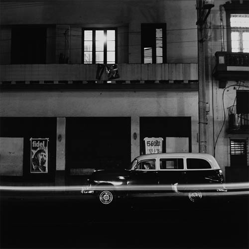 A long exposure photograph of the exterior of a very old apartment building. The face of the building fills the entire frame. The lights are on in the apartment and it is beaming out of the windows and shining through the louvers of the shutters. There is a balcony in the middle of the frame, a Cuban flag and photo of Fidel Castro hang on the edge. On the ground level of the building a poster is stuck to the wall, the poster features an illustration of Fidel Castro’s face with a star behind the head, the words "Fidel Cuba" are written at the top. In front of the building a 1950s vintage sports utility vehicle is parked. A man sits in the driver’s seat, his face is illuminated by a cellphone screen, his face is slightly blurry from movement during the long exposure of the photograph. Light streaks span the entire photograph in front of the vehicle, left behind by a passing car during the exposure of the photo.