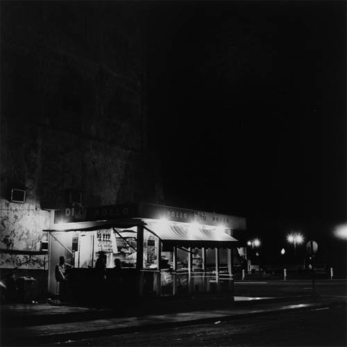 A long exposure photograph of a street food stand on the corner of two streets. The photograph is framed at an angle so two sides of the stand can be seen. The name "Pollo Ditu" is written on the top of the stand. On one side patrons can be seen sitting, two are dark and obscured by motion blur, a third can be seen in more still detail illuminated by the lights from inside the stand. Inside the stand cooks can be seen tending to their grills. Behind the stand the faint details of a large wall can be seen. The wall extends to the top of the frame. Street lights can be seen in the distance illuminating the road on the far side of the stand.