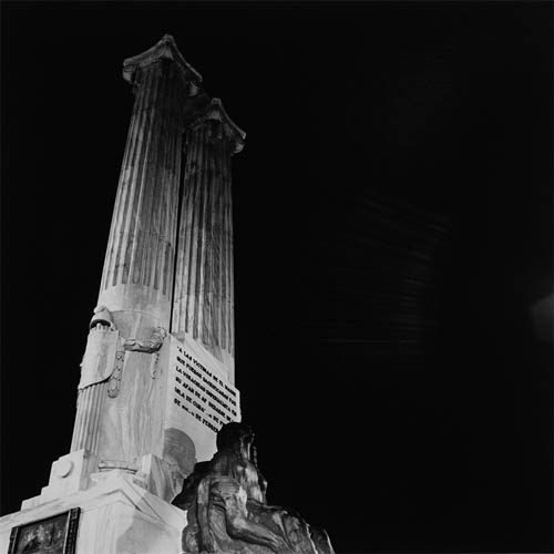 A long exposure photograph of a monument with two Ionic columns at the base is a statue of a woman. The woman is looking toward the sky, on her side is the skeletal body of a person, its hard to tell if this is a man or a woman. Above the statue is a plaque with the words "A las víctimas de El Maine que fueron sacrificadas por la voracidad imperialista en su afán de apoderarse de la isla de Cuba. Febrero 1898 – Febrero 1961". The columns and statue are fully lit, beyond is a completely black sky.