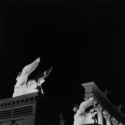 A long exposure photograph featuring two statues of griffins at an angle. The griffins are perched on ornate pillars. They appear to be made of marble or something made to resemble white marble. They are well lit against the dark sky. in the near distance a higher ledge supported by several Ionic columns can be seen. On the ledge statues of two angels are visible playing long trumpets.