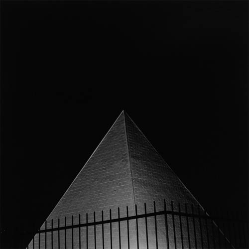 A long exposure photograph of a pyramid made of rectangular white tiles. The pyramid is brightly lit against a dark sky. In front of the pyramid the dark silhouette of an iron fence can be seen. As the fence extends beyond the pyramid, you can very slightly still see the dark fence against the black sky. The size and purpose of the pyramid is a mystery.