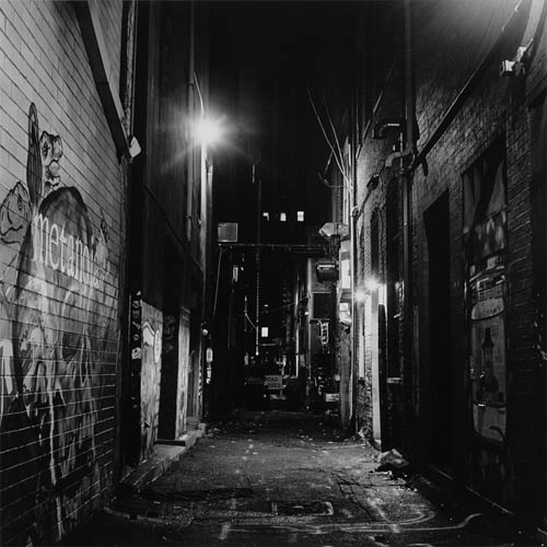 A long exposure photograph of the middle of an alley, the path is narrow with buildings tight on each side of the frame. The photo is a bit claustrophobic and uncomfortable. There is graffiti on both sides of the alley most of it is hard to make out at the extreme angle, but on the left side of the frame a mural of a human skull can be seen. There are several lights illuminating the alley into the distance. In the middle of the path in the background a sign can be seen that reads "Road Closed" adding to the uninviting feeling of the place.