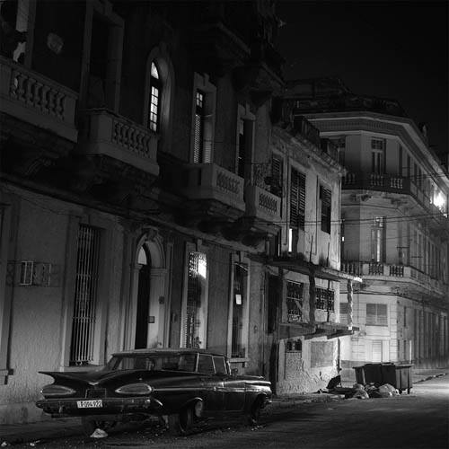 A long exposure photograph looking down a street at an angle. Old buildings can be seen lining the street. A light is shining from out of the frame illuminating the buildings at an angle. The buildings have ornate balconies with windows. Some of the lights are on in the windows. In the foreground parked in front of the nearest building is a vintage 1950s 4 door car with horizontal fins and horizontal tear drop shaped taillights. In front of the car is a dumpster full of garbage. There is a quiet yet mysterious mood to the scene.