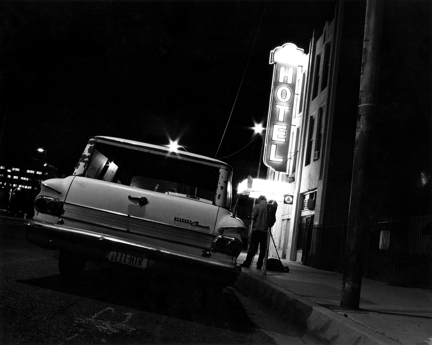 Behind the scenes of the artist Brian Seek taking a long exposure photograph of the New Windsor Hotel. The hotel is in the background, the photographer with tripod is in the middle ground, and in the foreground is Brian Seek's old station wagon.